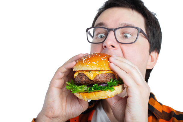 Taze bir Burger tutan gözlüklü genç bir adam. Çok aç bir öğrenci fast food yiyor. Sıcak yararlı yemek. Oburluk ve sağlıksız beslenme kavramı. Metin için kopya yer ile. İzole. - Fotoğraf, Görsel