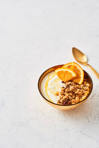Bowl of homemade granola with yogurt, honey and fresh orange on white abstract background. Healthy breakfast. Golden utensils. Top view. Copy space for text. - Photo, image
