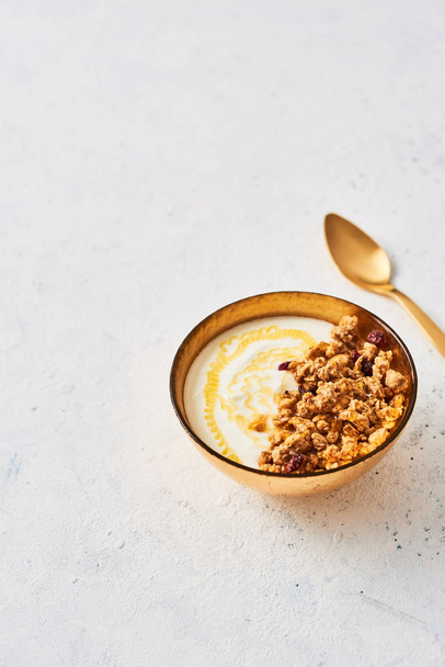 Bowl of homemade granola with yogurt and honey on white abstract background. Healthy breakfast. Golden utensils. Top view. Copy space for text. - Photo, image