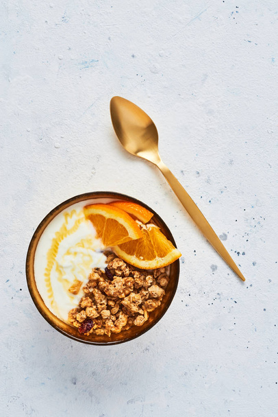 Bowl of homemade granola with yogurt, honey and fresh orange on white abstract background. Healthy breakfast. Golden utensils. Top view. Copy space for text. - Φωτογραφία, εικόνα