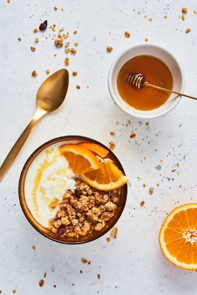 Bowl of homemade granola with yogurt, honey and fresh orange on white abstract background. Healthy breakfast. Golden utensils. Top view. Copy space for text. - Photo, Image