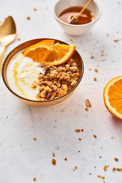 Bowl of homemade granola with yogurt, honey and fresh orange on white abstract background. Healthy breakfast. Golden utensils. Top view. Copy space for text. - Foto, Imagem