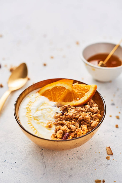 Bowl of homemade granola with yogurt, honey and fresh orange on white abstract background. Healthy breakfast. Golden utensils. Top view. Copy space for text. - Photo, Image