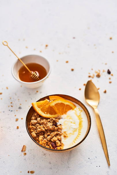 Bowl of homemade granola with yogurt, honey and fresh orange on white abstract background. Healthy breakfast. Golden utensils. Top view. Copy space for text. - Foto, imagen
