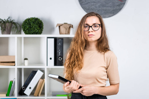 Portréja egy fiatal kaukázusi nő hosszú haj és a szemüveg a súrlódás irodát, egy office szobában egy íróasztal közelében áll. Egyetemi hallgató lány. A témakör az ember tanulási. - Fotó, kép