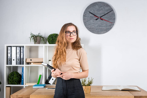 Retrato de uma jovem caucasiana com cabelos longos e óculos para fricção fica em um escritório dentro de uma sala de escritório perto de uma mesa. Uma estudante universitária. Tópico homem aprendizagem
. - Foto, Imagem