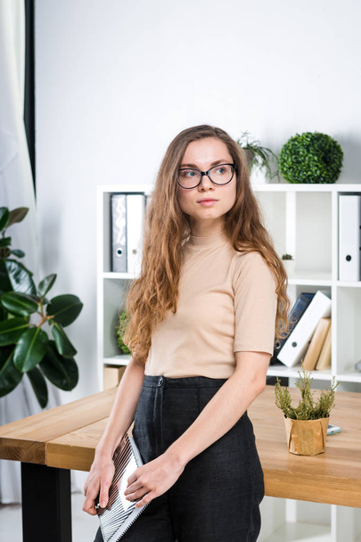 Portrait of a young Caucasian woman with long hair and glasses for friction stands in an office inside an office room near a desk. University student girl. Topic man learning. - Foto, Imagen