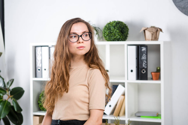 Portrait of a young Caucasian woman with long hair and glasses for friction stands in an office inside an office room near a desk. University student girl. Topic man learning. - Foto, afbeelding