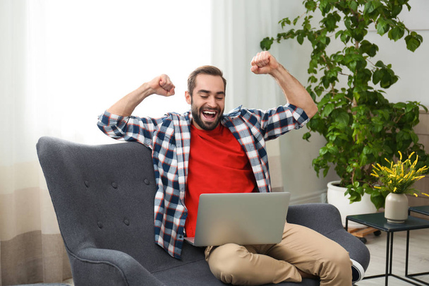Emotional young man with laptop celebrating victory on sofa at home - Foto, Bild