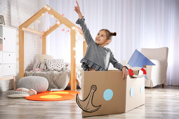 Cute little girl playing with cardboard boat in bedroom - Photo, image
