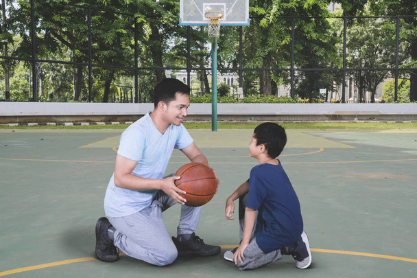 Picture of young man teaching his son to play basketball while kneeling in the basketball court - Foto, Imagem