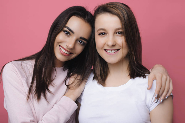 Nahaufnahme Porträt zweier erstaunlicher Freundinnen, die sich umarmen und lächelnd in die Kamera an der rosafarbenen Wand schauen. zwei schöne Frauen posieren mit einem Lächeln im Gesicht vor der Kamera. - Foto, Bild