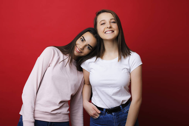 Two amazing young girls looking at the camera smiling while one is leaning her head on another's shoulder against red wall. Girlfriends having fun together laughing. - Фото, зображення