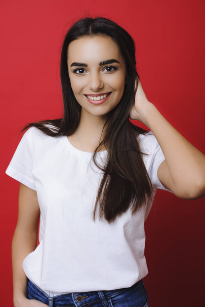 Adorable brunette girl dressed in white shirt and blue jeans looking into camera smiling while touching her hair naturally against a red background. - Fotó, kép