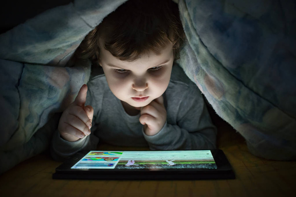 Mała dziewczynka oglądając jej tabletki w łóżku. Oświetlona twarz dziecka z ekranu urządzenia. Dziecko ubrane z piżamy pod kołdrą przytrzymaj tabletkę. Pora nocna. - Zdjęcie, obraz