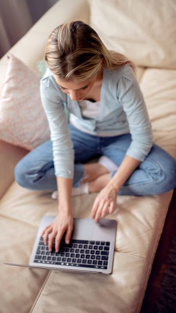 Дом, технологии, коммуникации и люди - улыбающаяся женщина, сидящая на диване и сидящая на ноутбуке
. - Фото, изображение