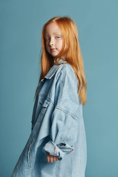 Beautiful redheaded baby girl with long hair in a big blue long denim jacket. Fashion clothing, spring portrait of red girl on blue background. Big beautiful eyes of a young girl, a vivid portrait - Foto, Bild
