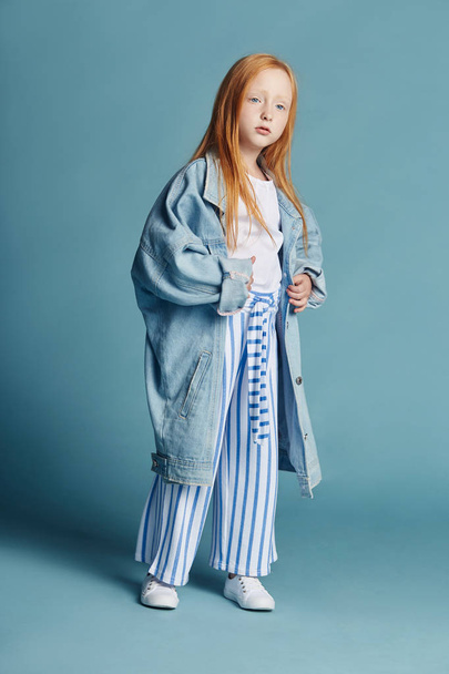 Beautiful redheaded baby girl with long hair in a big blue long denim jacket. Fashion clothing, spring portrait of red girl on blue background. Big beautiful eyes of a young girl, a vivid portrait - Foto, imagen