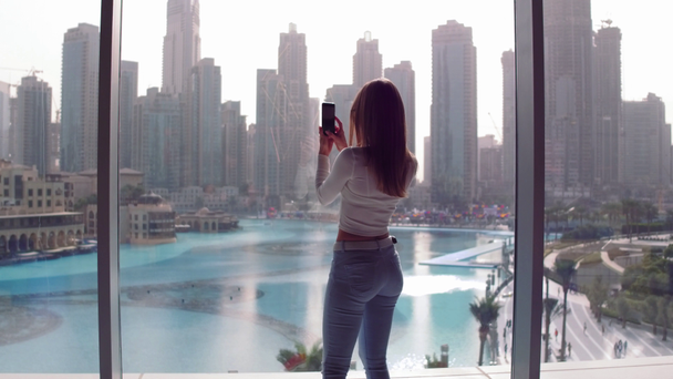 Meisje permanent in de buurt van het venster van een hoog gebouw in Dubai city en kijkt neer op het meer en de wolkenkrabbers. - Video