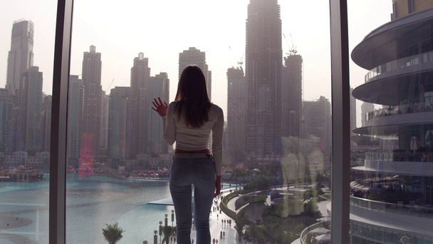 Young woman coming to the window and looking and looking down to the lake and skyscrapers at the subset. - Footage, Video