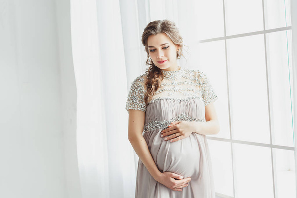 灰色のドレスを着て、白いスタジオでポーズをとる上品な美しい妊娠女性の肖像画。妊娠中の女性は、彼女の腹に触れます。美しい、健康と幸せな妊娠の概念 - 写真・画像