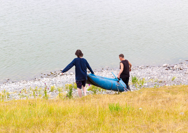 Sayanogorsk, Khakassia, Rusia - 17 de julio de 2018: Hombre adulto y un adolescente juntos llevan un bote de goma y remos para pescar en el lago. Vacaciones turísticas o caminata en una sombría mañana de verano
 - Foto, imagen