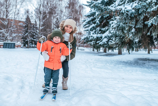 Young happy mother, a woman holds a boy 2-5 years old son, learns to ski. In winter in park outside. Caring for the baby, raising a child. In a warm hat and jacket. Free space for text. - Fotó, kép
