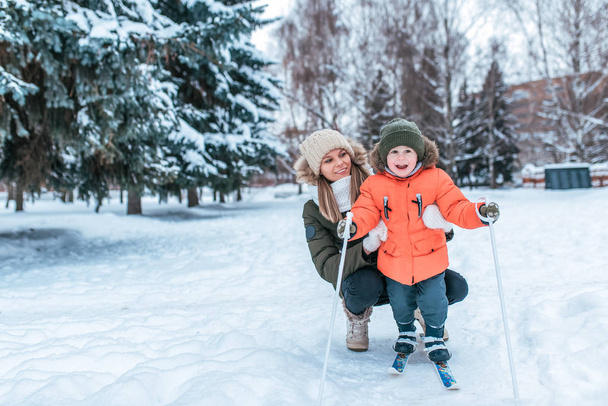 Young happy mother, a woman holds a boy 3-5 years old son, learns to ski. In winter in park outside. Free space for text. Caring for the baby, raising a child. In a warm hat and jacket. - Fotoğraf, Görsel