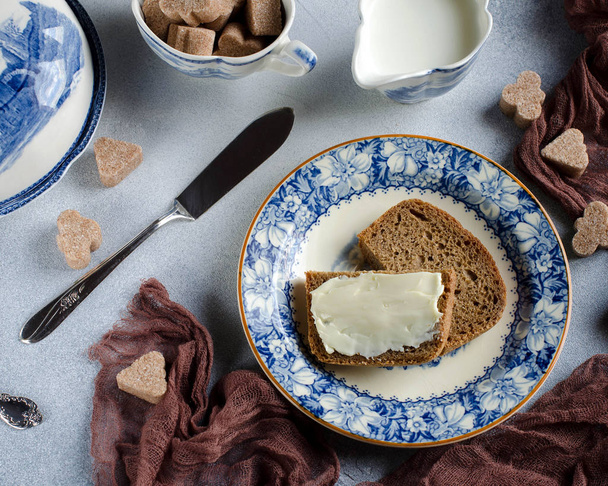 Breakfast on the table-top view. Rye bread with butter on a plate. Cane sugar. Beautiful Breakfast serving. Blue plates on grey background. Vintage Cutlery.  Brown runner. - 写真・画像