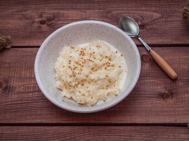 Colazione leggera di porridge di latte di riso in una tazza bianca profonda, cosparsa di semi di lino. Un piatto su un vassoio marrone alla moda. Accanto al cucchiaio con una maniglia in legno
. - Foto, immagini
