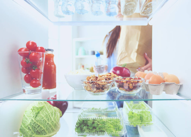 Portrait of female standing near open fridge full of healthy food, vegetables and fruits. - Foto, afbeelding