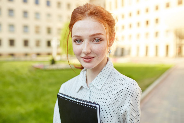Cheerful attractive young woman with freckles and notebooks standing and smiling - Foto, Bild