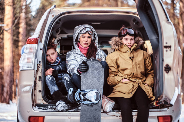 Una hermosa mujer y sus hijos vestidos con ropa de abrigo sentados en el maletero de un coche
 - Foto, imagen