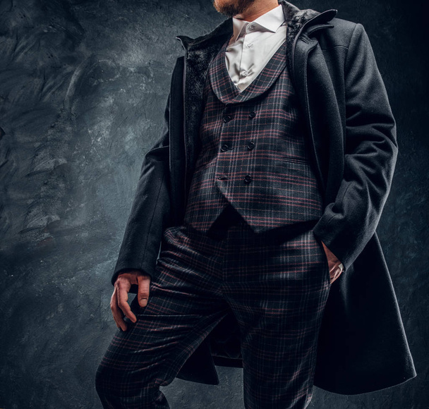 A stylish man dressed in an elegant suit and coat posing with a hand in a pocket against a dark textured wall - Foto, imagen