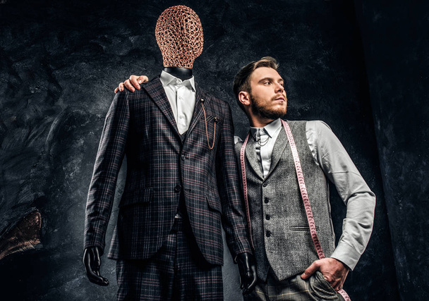 Un joven diseñador de moda confiado posando junto a un maniquí en traje exclusivo hecho a medida para hombre en un estudio de sastre oscuro
  - Foto, imagen
