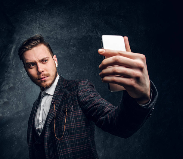 Magabiztos fiatal stílusos ember öltözött egy elegáns ruha, figyelembe selfie okostelefon-egy sötét stúdióban - Fotó, kép