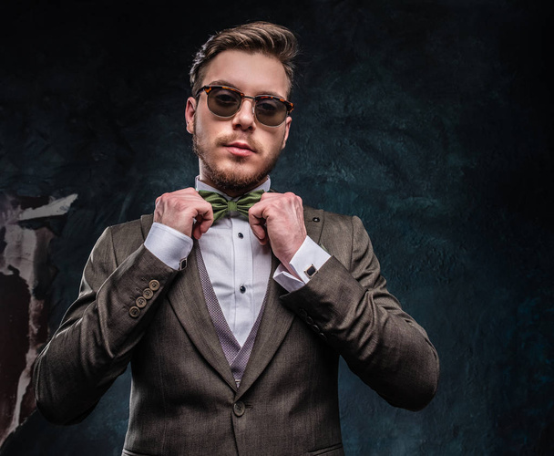 Стильный молодой человек, одетый в элегантный костюм, правит галстук-бабочку о темную текстуру стены.
 - Фото, изображение