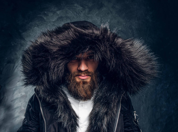 Πορτρέτο του γενειοφόρος άνδρας σε ένα παλτό με κουκούλα γούνα σε ένα σκοτεινό υφή τοίχο - Φωτογραφία, εικόνα