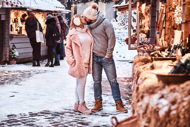 Una atractiva pareja enamorada usando ropa de abrigo elegante cogida de la mano y besándose mientras está de pie en la feria de invierno en época de Navidad
. - Foto, Imagen