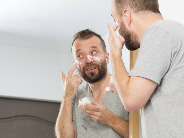 Happy funny adult guy applying moisturizer cream for male skin care. Man in front of bathroom mirror with anty aging lotion on face. - Фото, зображення