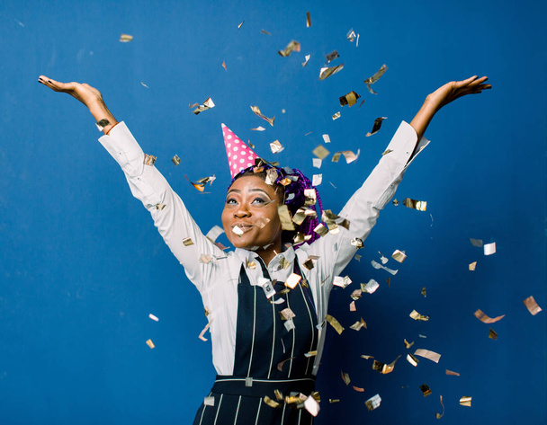 beautiful young black woman celebrating birthday or new year and chrismas party while blowing confetti decorations to camera isolated over blue background - Фото, изображение