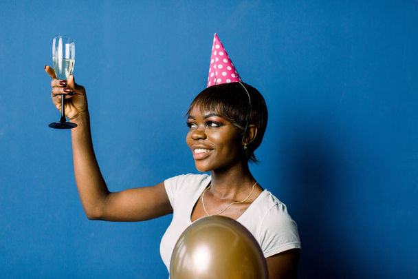 Portrait of beautiful birthday african girl resting at party. Pretty smiling african girl holding glass with champagne and air balloons posing on blue background - Foto, imagen