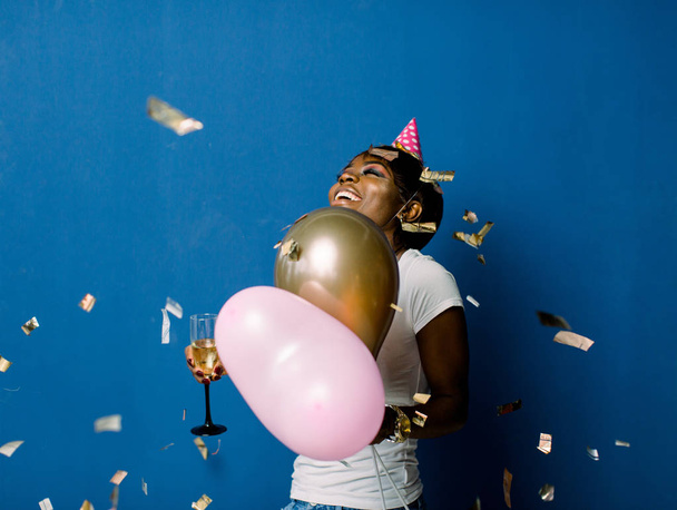 Glad pretty african woman in white t-shirt happy dancing and throwing out confetti, celebrating birthday. Indoor photo of pretty black lady holding champagne and balloons with pleased face expression - Photo, Image