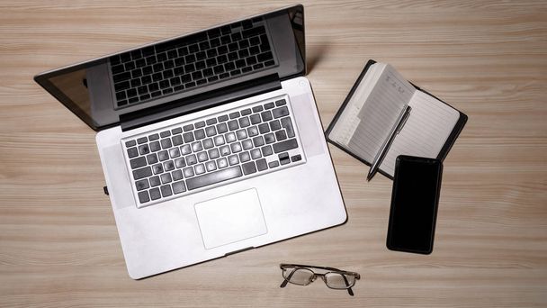 Desk with laptop, eyeglasses, notepad, smartphone, and pen on a wooden table. Top view with copy space. Flat lay - image - Фото, изображение
