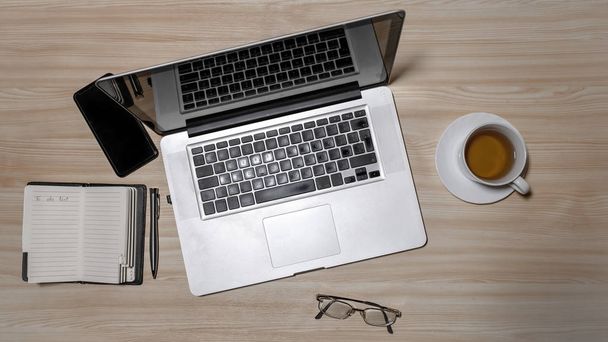 Desk with laptop, eyeglasses, notepad, smartphone, pen and a cup of tea on a wooden table. Top view with copy space. Flat lay - image - Фото, изображение