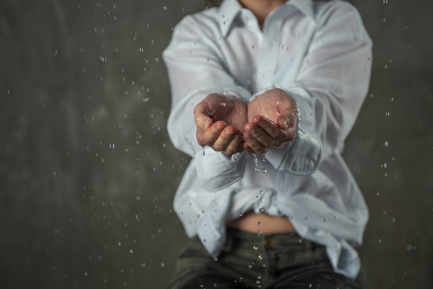 drops of water fall and flow out of women's hands, the girl dressed in a white shirt stands on a gray wall - Foto, Imagen