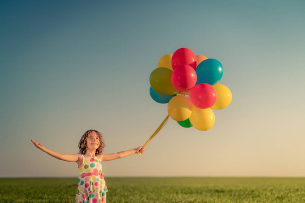 Niño feliz jugando con globos multicolores brillantes al aire libre. Niño divirtiéndose en el campo de primavera verde contra el fondo azul del cielo. Concepto de estilo de vida saludable y activo - Foto, Imagen