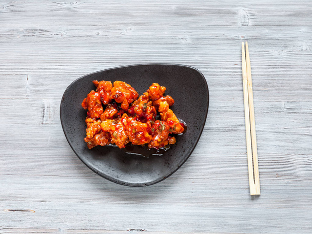 Korean Chinese cuisine - top view of Kkanpunggi spicy fried Chicken pieces with vegetables in sweet and sour sauce on black plate with chopsticks on gray table - Photo, Image