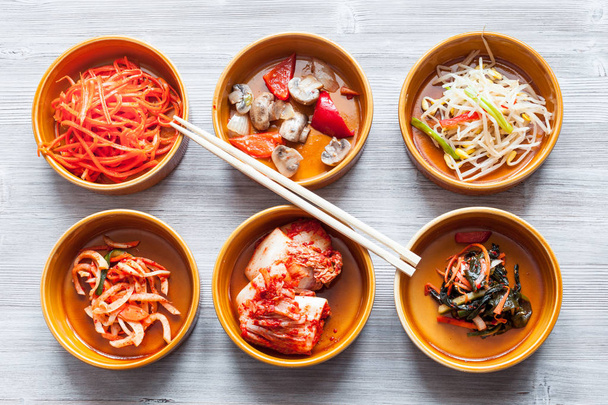 корейская кухня - палочки для еды над различными закусками (Банчан или Панчан) в керамических мисках на сером столе
 - Фото, изображение