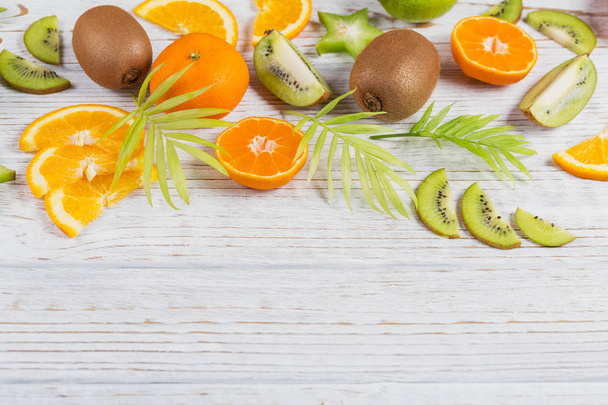 Green palm leaves and fresh tropical slices of fruits on white wooden table. Juicy oranges, tangerines, limes, kiwis, carambolas. Summer fruit background. Flat lay, top view, copy space - Photo, Image